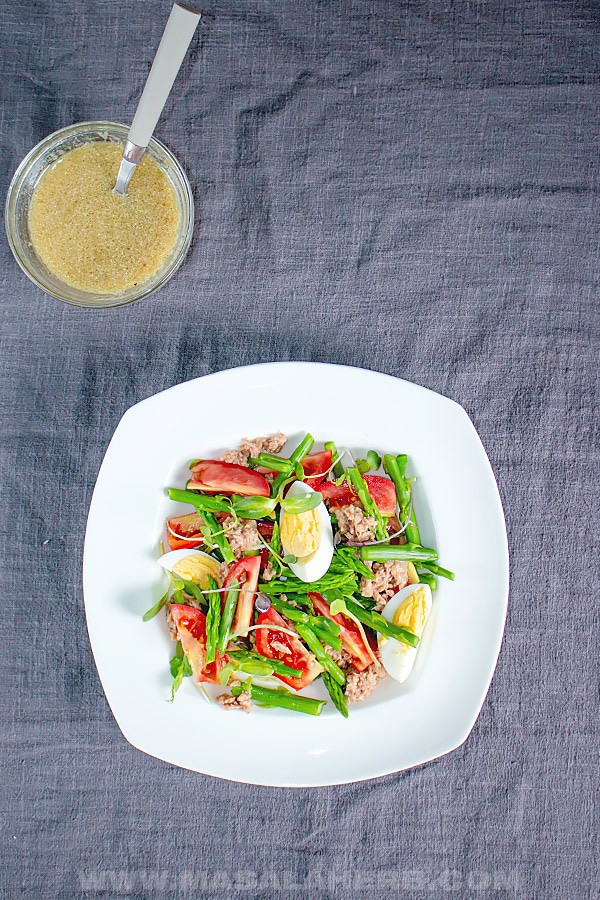 Overhead view of Tuna Asparagus Salad with Microgreens with eggs and tomatoes on white place with cup of dressing on side 