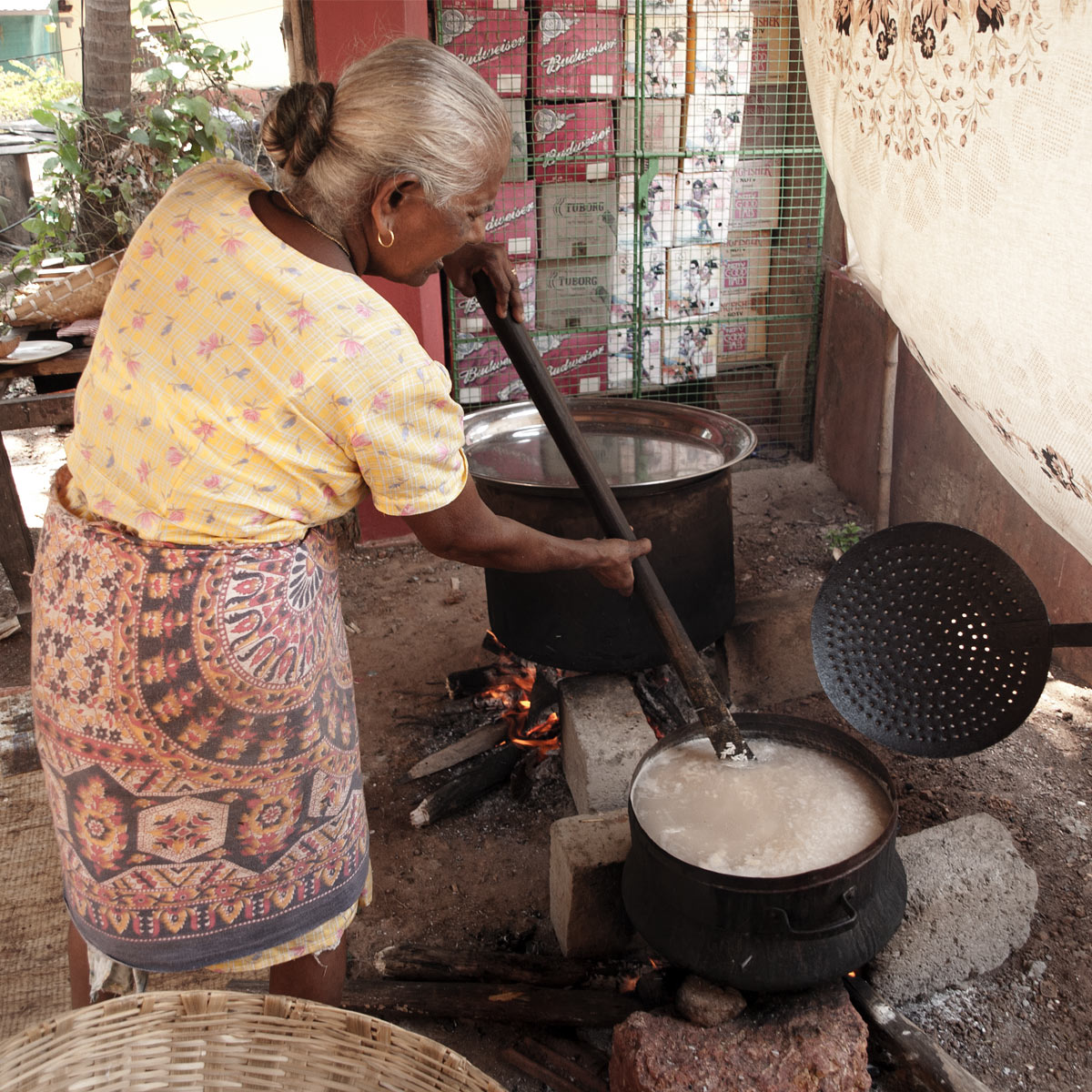 Goan lady stirring a large ladder in a large pot filled with cooking rice over an open wood fire place