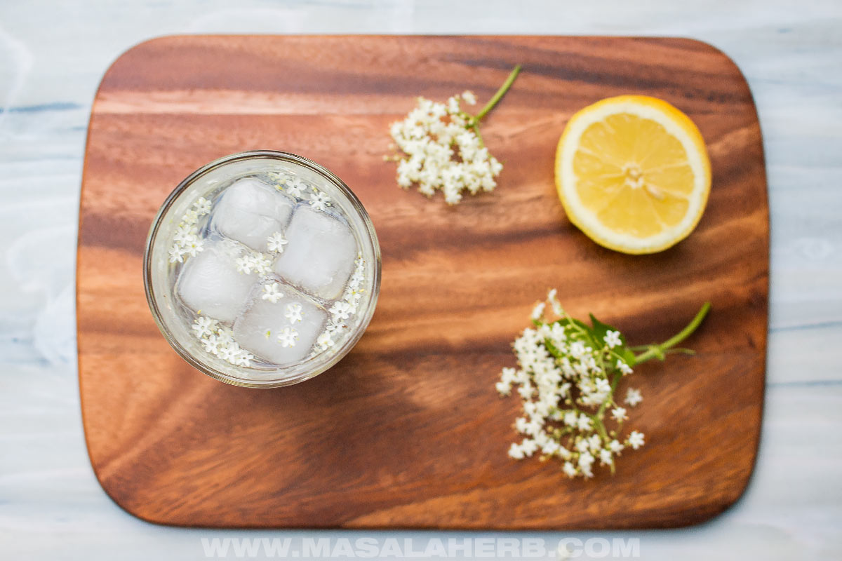 elderflower cocktail drink with fresh elderflower blossom bunch and lemon half on a mango cutting board top down view